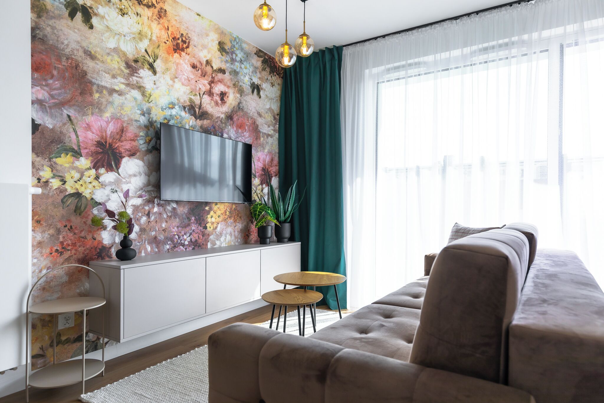Una sala moderna con un televisor de pantalla plana grande montado en una pared con un mural floral, un sofá gris, dos mesitas redondas y una cortina verde.