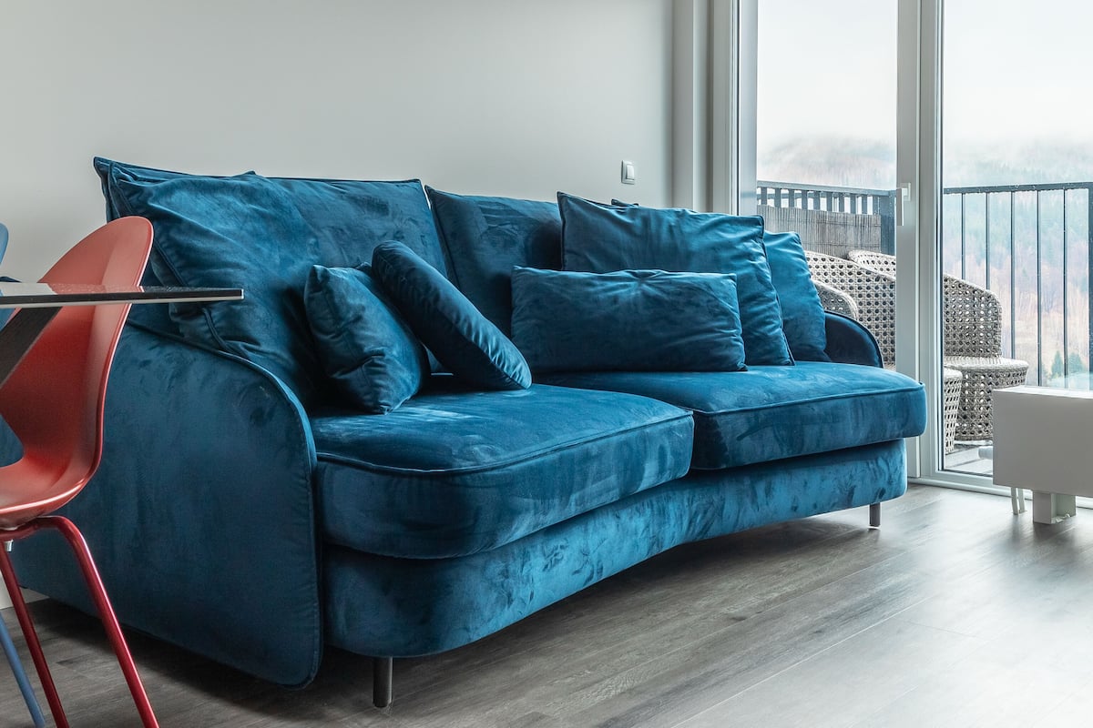 Un salon moderne avec un canapé en velours bleu, un fauteuil rouge et un balcon offrant une vue dégagée.
