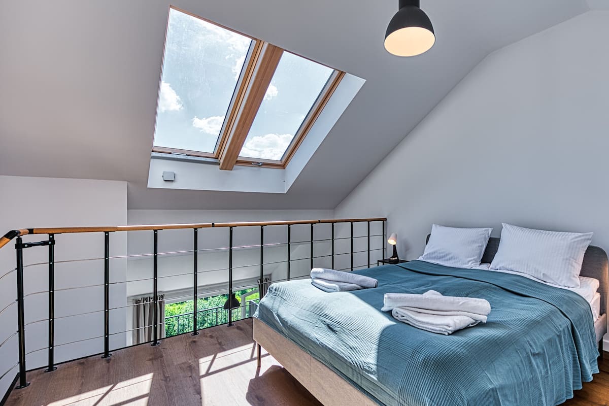 Une chambre avec un puits de lumière, un lit recouvert d’une couverture bleue, une table de chevet avec une lampe, et une vue sur la verdure à l’extérieur.
