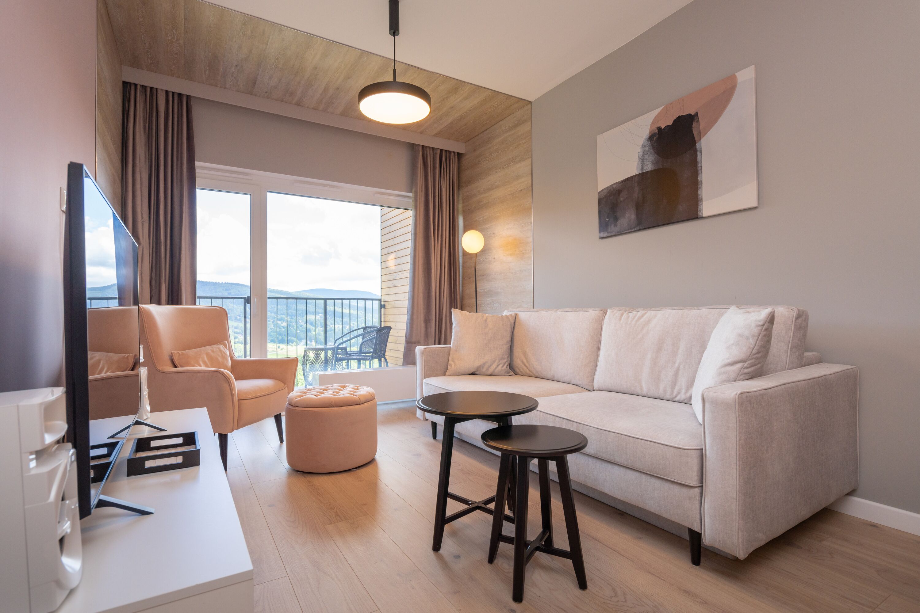 A modern living room with a sofa, a coffee table, a TV, and a chair. There is a balcony with a view of the mountains.