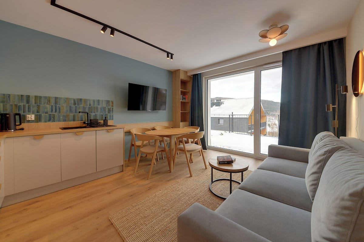 Un salon moderne avec un canapé gris, une table à manger en bois avec des chaises, un coin cuisine équipé d’une machine à café et une grande fenêtre donnant sur un paysage enneigé.