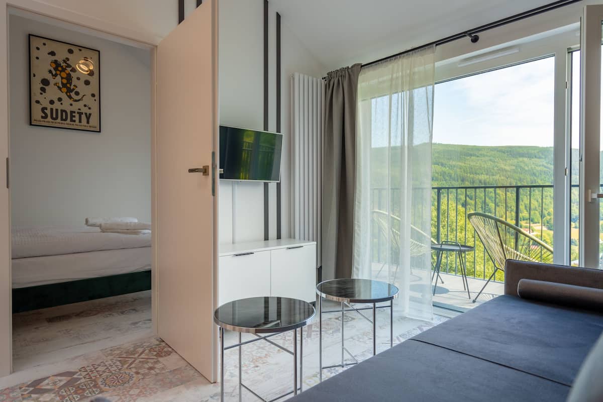 Une chambre d’hôtel moderne équipée d’un lit, d’une télévision, d’un balcon avec une chaise et offrant une vue sur la forêt.