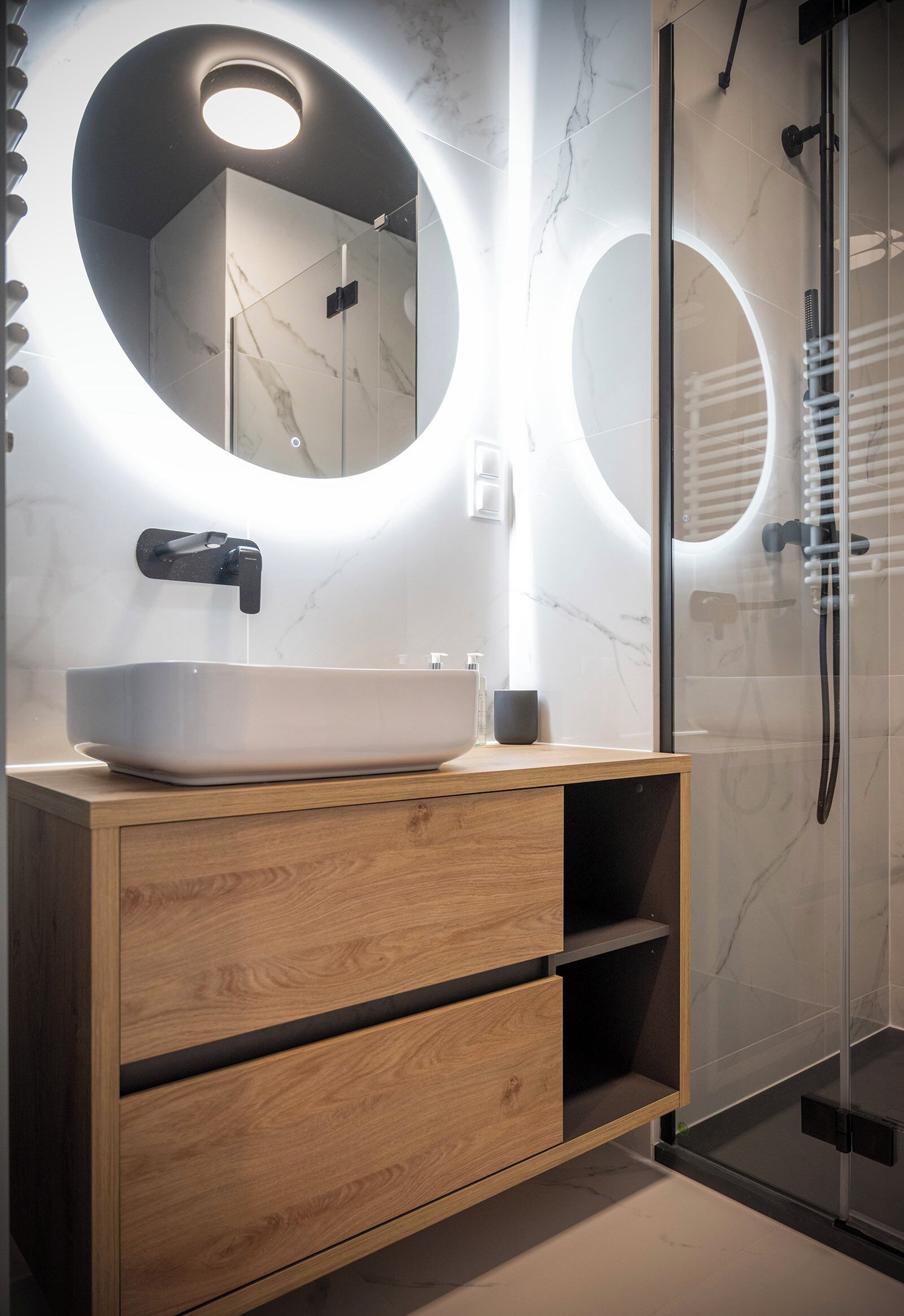 A modern bathroom with a large oval mirror, a vessel sink, wooden cabinetry, and a glass-enclosed shower.