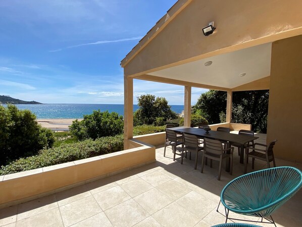 A covered outdoor area with a table and chairs, overlooking a beach and the sea.