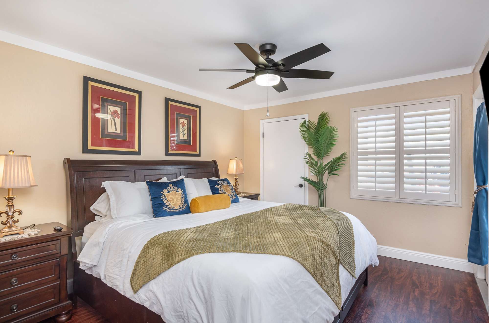 Une chambre à coucher comprenant un lit, une table de chevet, une lampe, un ventilateur de plafond, des tableaux encadrés accrochés au mur, une porte et une fenêtre avec des stores.