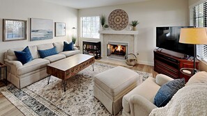 A living room with a sofa, ottoman, coffee table, fireplace, television, and bookshelves. - Brand NEW Remodel Single Level 2 Bd 2 Ba 2 Car Garage POOL UTC La Jolla Close to UCSD! (San Diego)