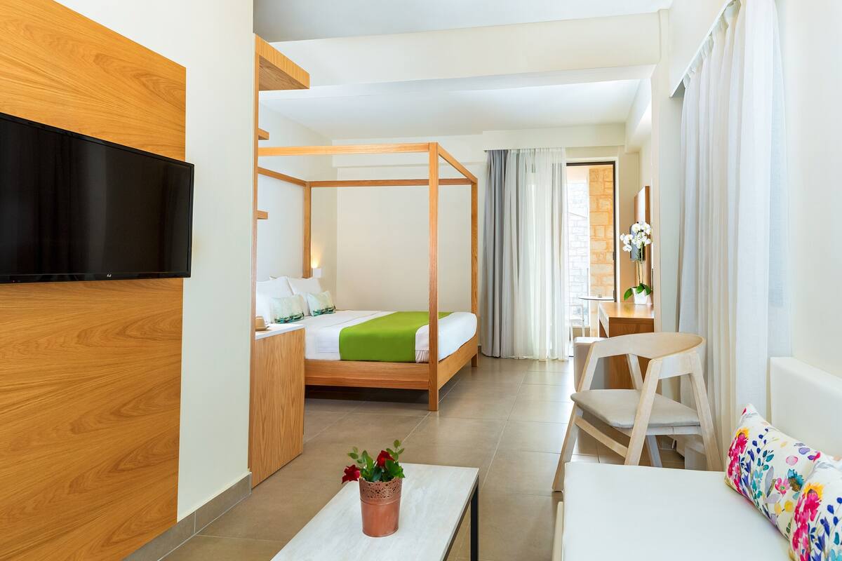 A modern hotel room with a wooden accent wall, a bed with a green bedspread, a desk with a chair, a TV mounted on the wall, and a small table with a plant.