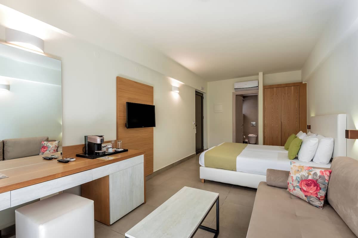 A modern hotel room with a wooden desk, a bed with a green and white duvet, a sofa with floral cushions, and a television mounted on the wall.