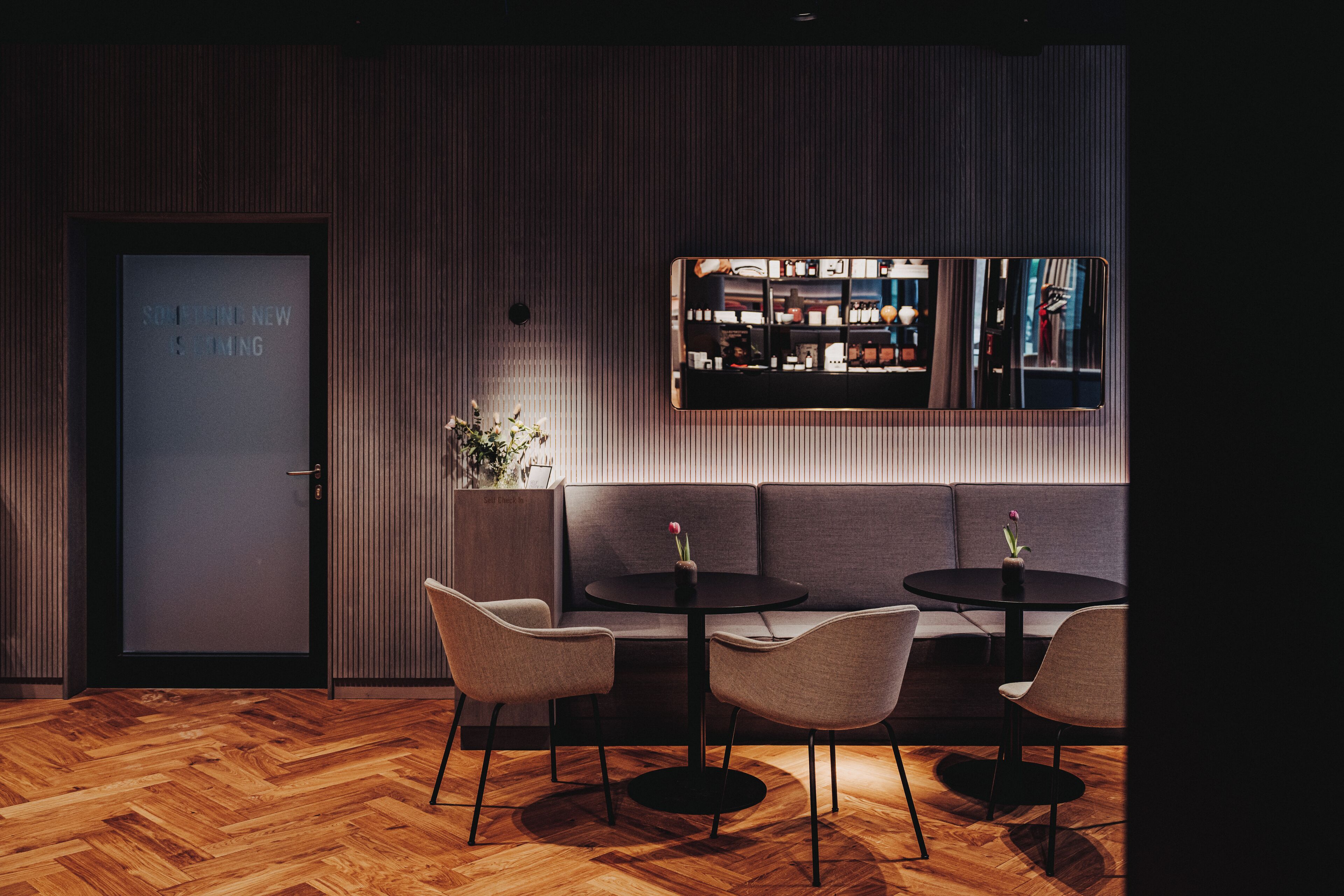 A modern interior with a wooden herringbone floor, a dark-colored sofa, two round tables with chairs, and a large mirror reflecting a bar area.