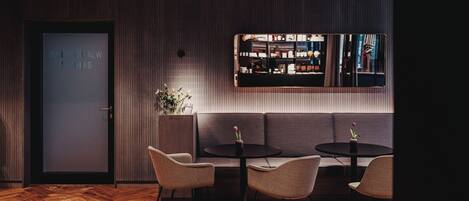 A modern interior with a wooden herringbone floor, a dark-colored sofa, two round tables with chairs, and a large mirror reflecting a bar area.