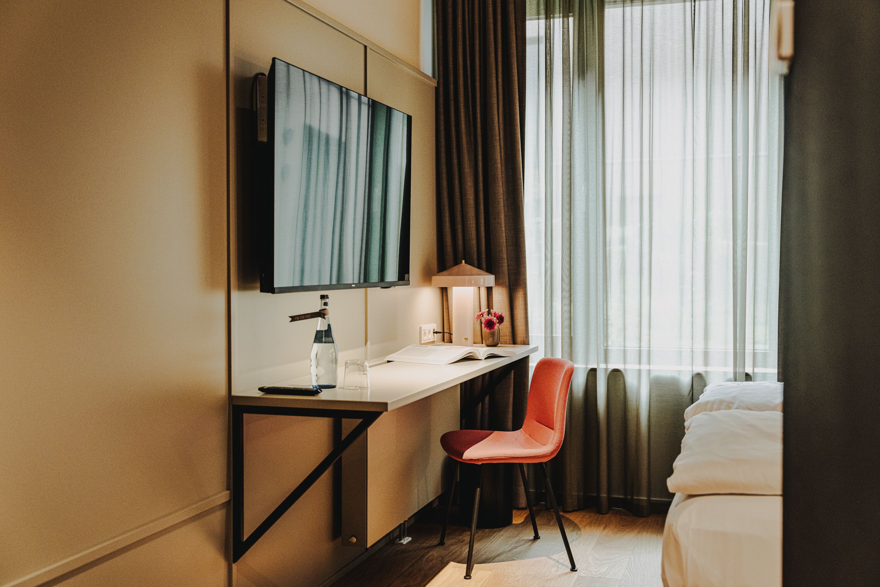 A hotel room with a bed, a desk with a chair, a TV, and a window with curtains.