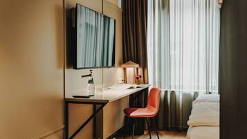 A hotel room with a bed, a desk with a chair, a TV, and a window with curtains.