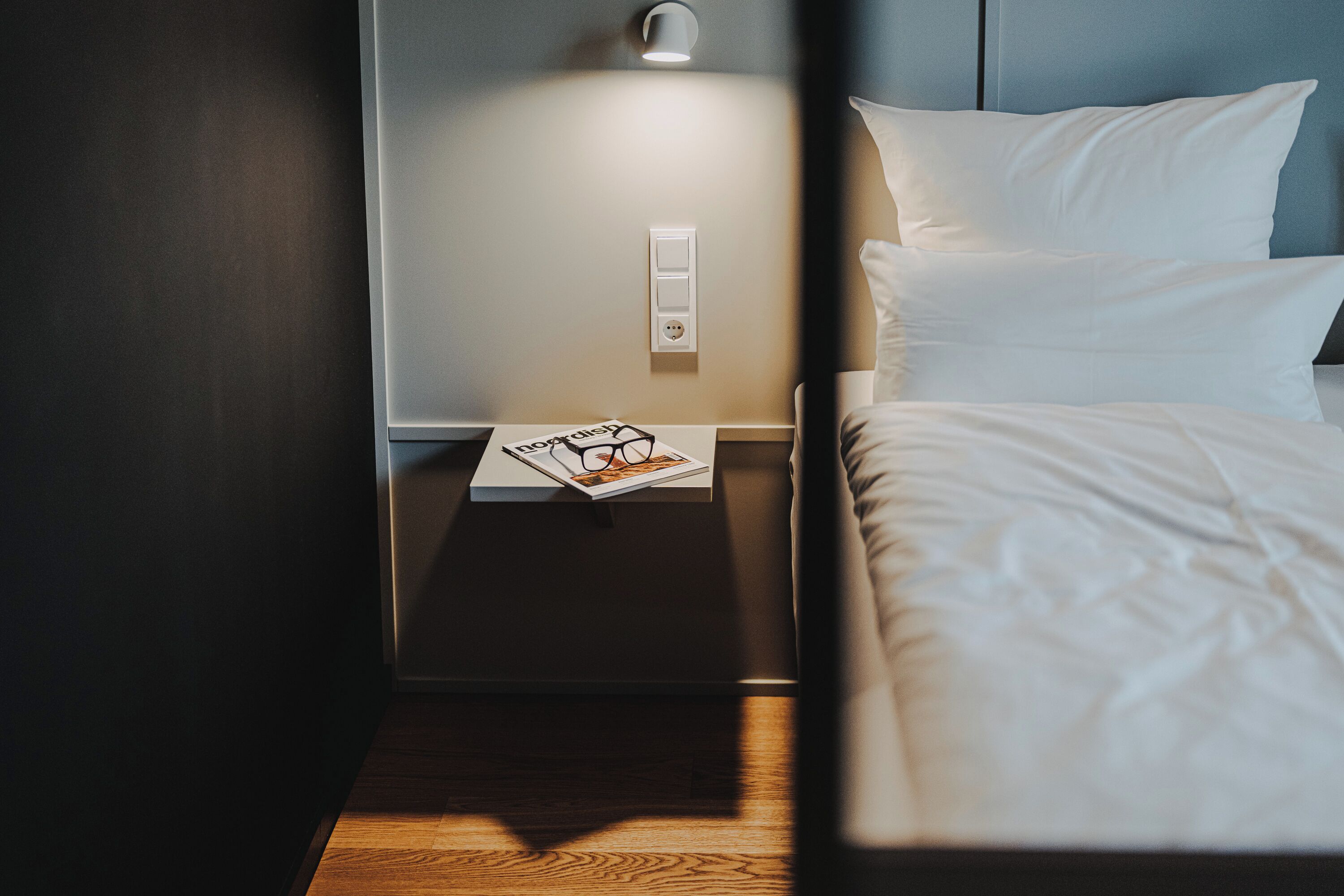 Een net opgemaakt bed met witte lakens, een nachtkastje met een lamp en een tijdschrift met brillen erop.
