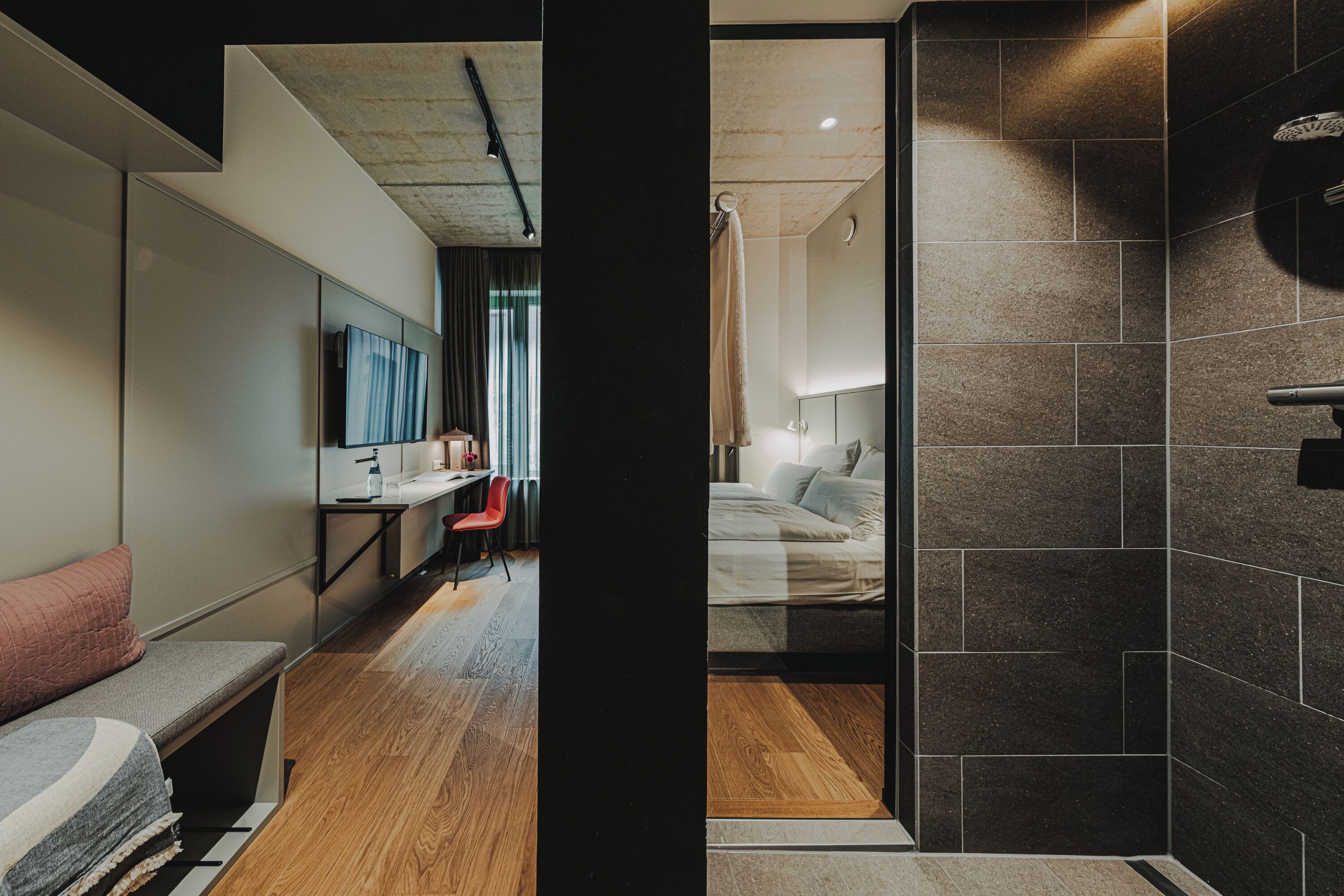 A modern hotel room with a wooden floor, a bed with white linens, a small desk with a chair, a large mirror, and a dark tiled shower area.