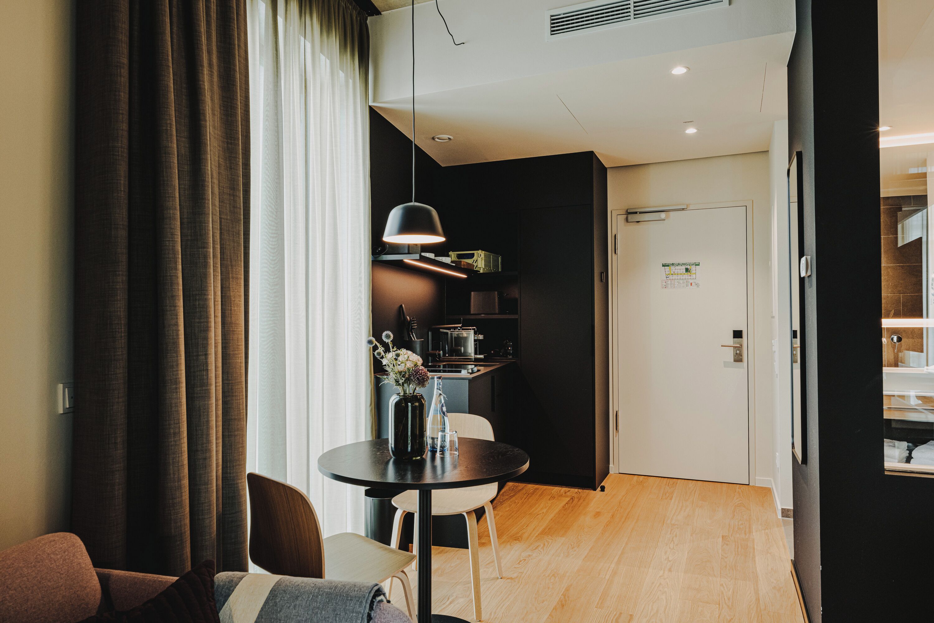A modern hotel room with a dining area, a kitchen, and a door leading to a bathroom.
