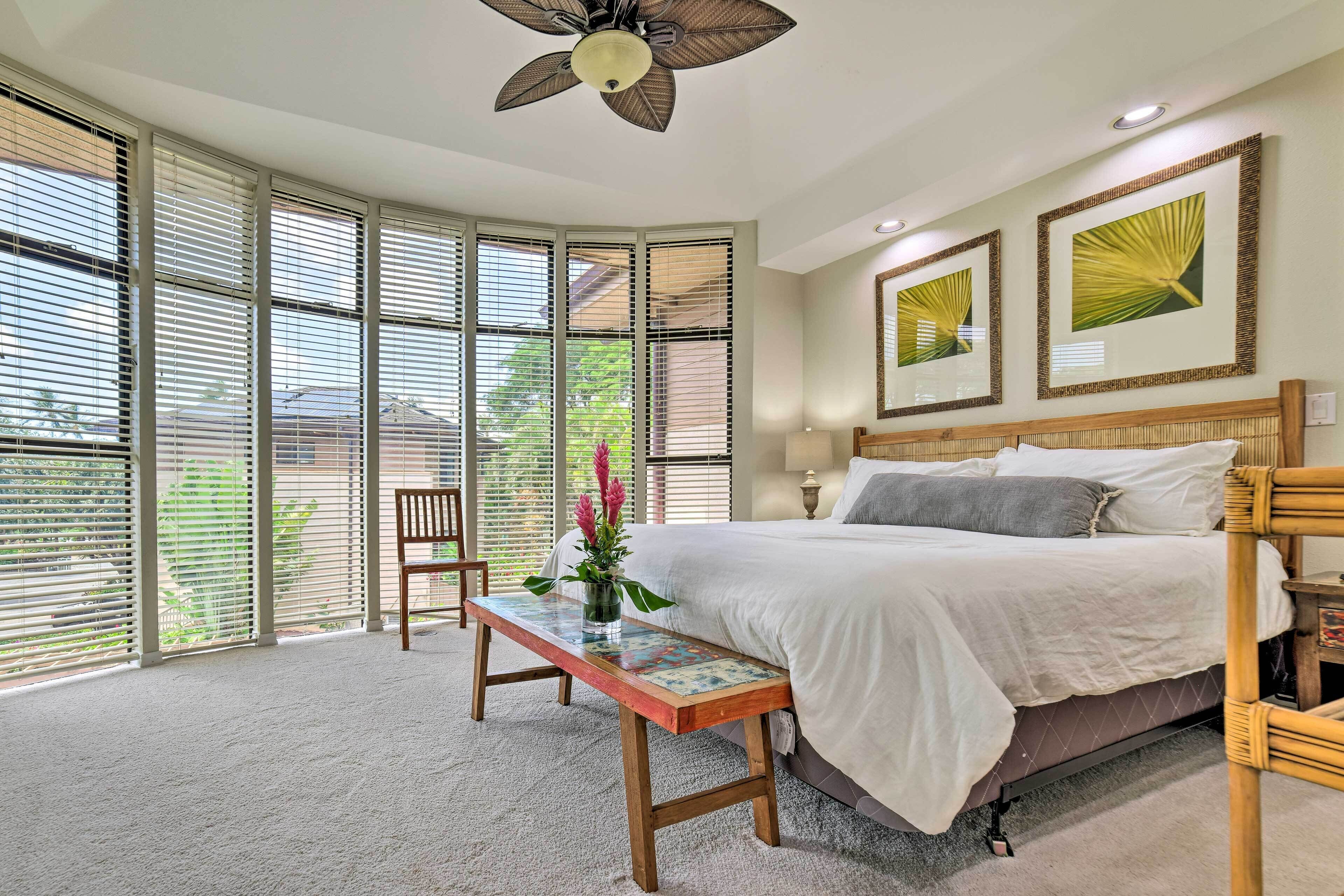 A bedroom with a bed, a wooden bench, a vase with flowers, and large windows with blinds.