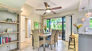 En matplats med flätade stolar, ett träbord och en takfläkt. Det finns också ett köksområde med en barstol och hängande lampor.