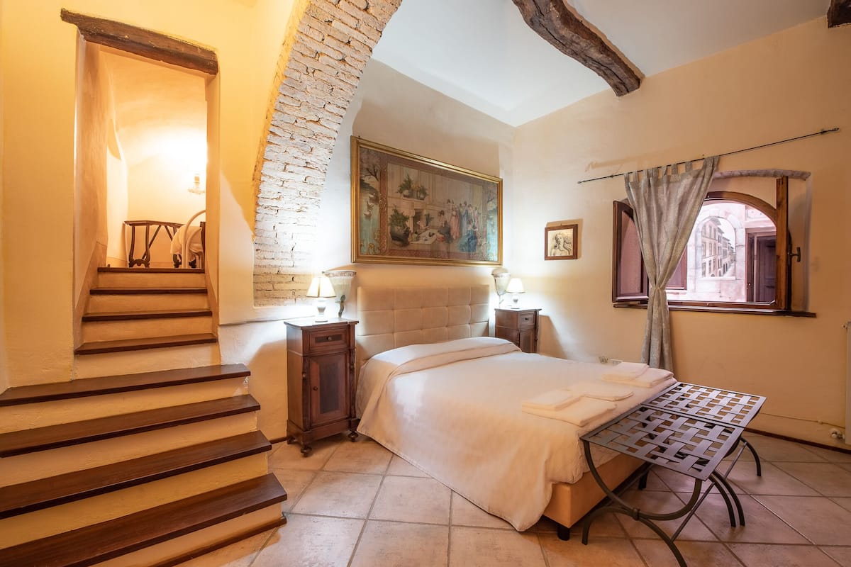 A bedroom with a bed, a nightstand, a lamp, a window with curtains, and a staircase.