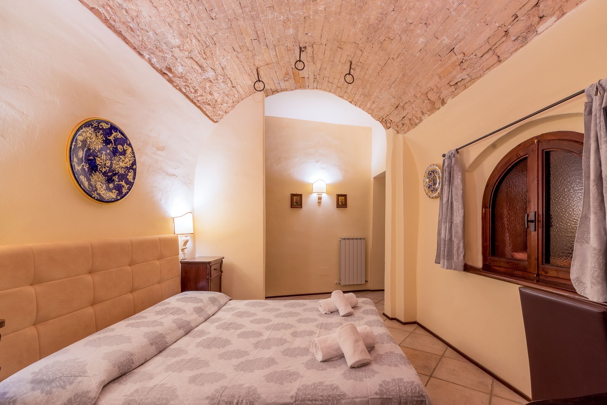 Une chambre avec un plafond en briques, un lit recouvert d’une couvre-lit à motifs floraux, une assiette décorative ronde accrochée au mur et une fenêtre avec des rideaux.