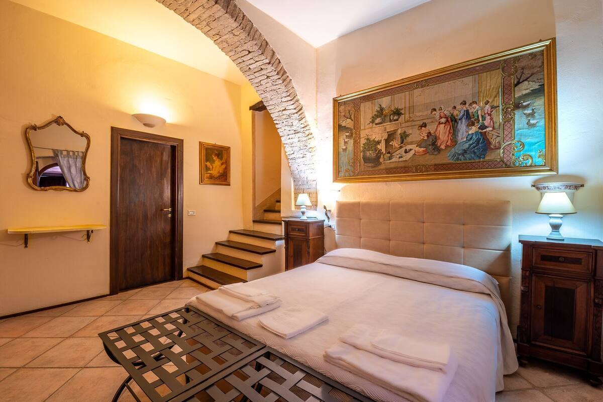A bedroom with a bed, a wooden headboard, a painting, a mirror, and a staircase.
