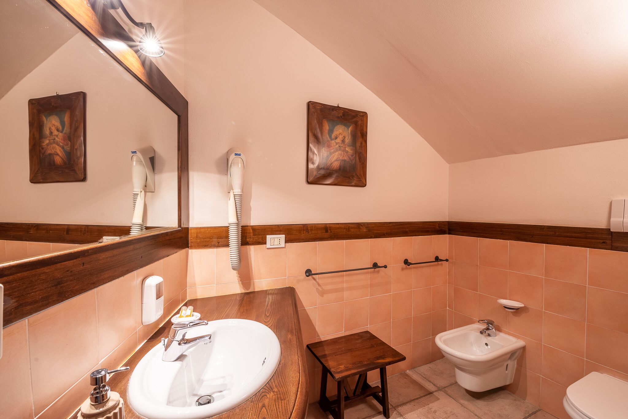 Un baño con tocador de madera, lavamanos, bidé y retrete. Hay dos toalleros en la pared y un espejo sobre el tocador.
