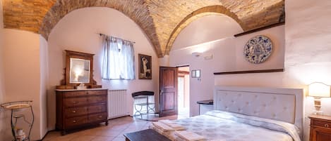 A bedroom with a brick ceiling, a bed with a headboard, a wooden dresser, a bench, and a wall-mounted plate.