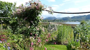 Un jardín con diversas plantas y flores, un puente colgante y una vista de montañas y un lago al fondo.
