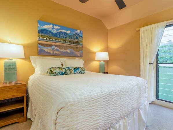A bedroom with a large bed, two bedside lamps, a painting on the wall, and a view through a window. - Spacious 3 Bedroom Ocean Front W/Lanai-Kaha Lani (Lihue)