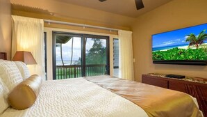Une chambre d’hôtel avec un lit, un ventilateur de plafond, une grande fenêtre avec des rideaux et un écran de télévision diffusant une scène de plage tropicale.