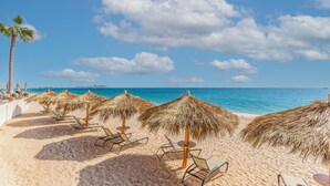 A beach with thatched umbrellas and lounge chairs, a palm tree, and a clear view of the sea.