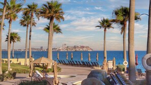 A seaside resort with palm trees, sun loungers, and a view of the ocean and cityscape.