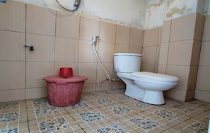 Ein kleines Badezimmer mit Toilette, einem roten Eimer und einem Duschschlauch.