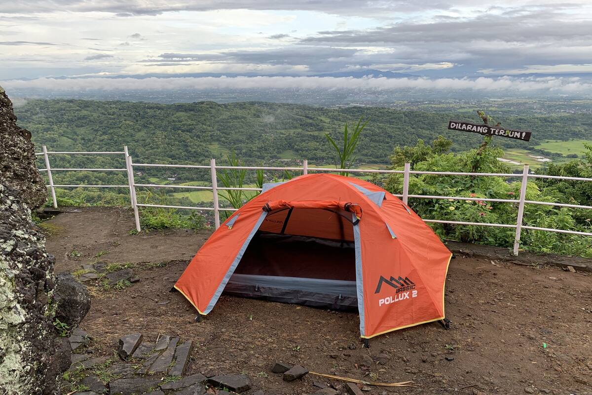 A red tent labeled "BASSIC POLLUX" on a rocky terrain with a view of a lush valley and a sign reading "DILARANG TERSUN!"