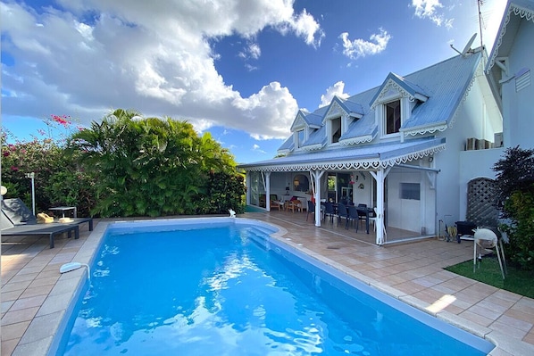 A swimming pool with a covered patio area, surrounded by lush greenery and a large house with a distinctive architectural style. - LA Maison DES Fées (Les Trois-Îlets)