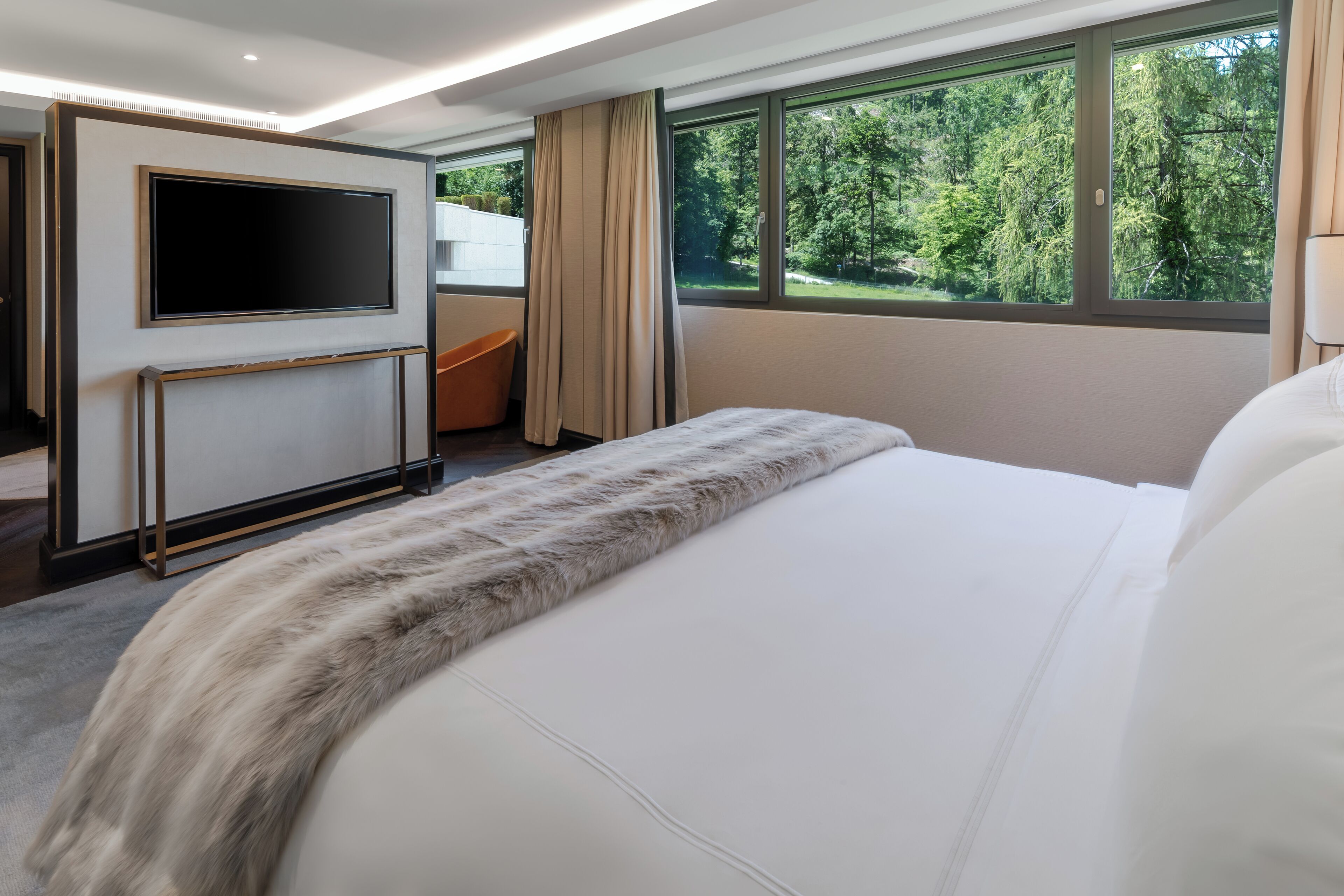 A modern hotel room with a large bed, a flat-screen TV mounted on the wall, a seating area with a chair and a small table, and large windows offering a view of the outdoors.
