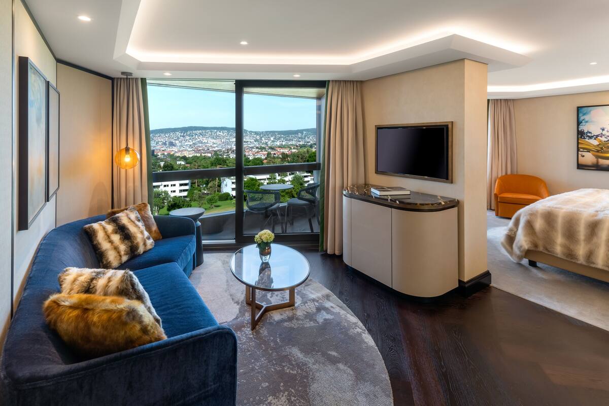 A modern hotel room with a sofa, a small round table, a TV, a bed, and a balcony with a city view.