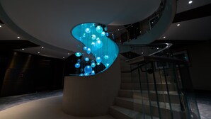 A modern interior with a curved staircase, glass railings, and a unique light installation. - FIVE Zurich - Luxury City Resort (Zürich)