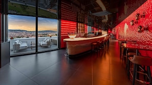 Un bar moderno con una pared roja de acento, amplios ventanales con vistas a la ciudad y una barra con taburetes.