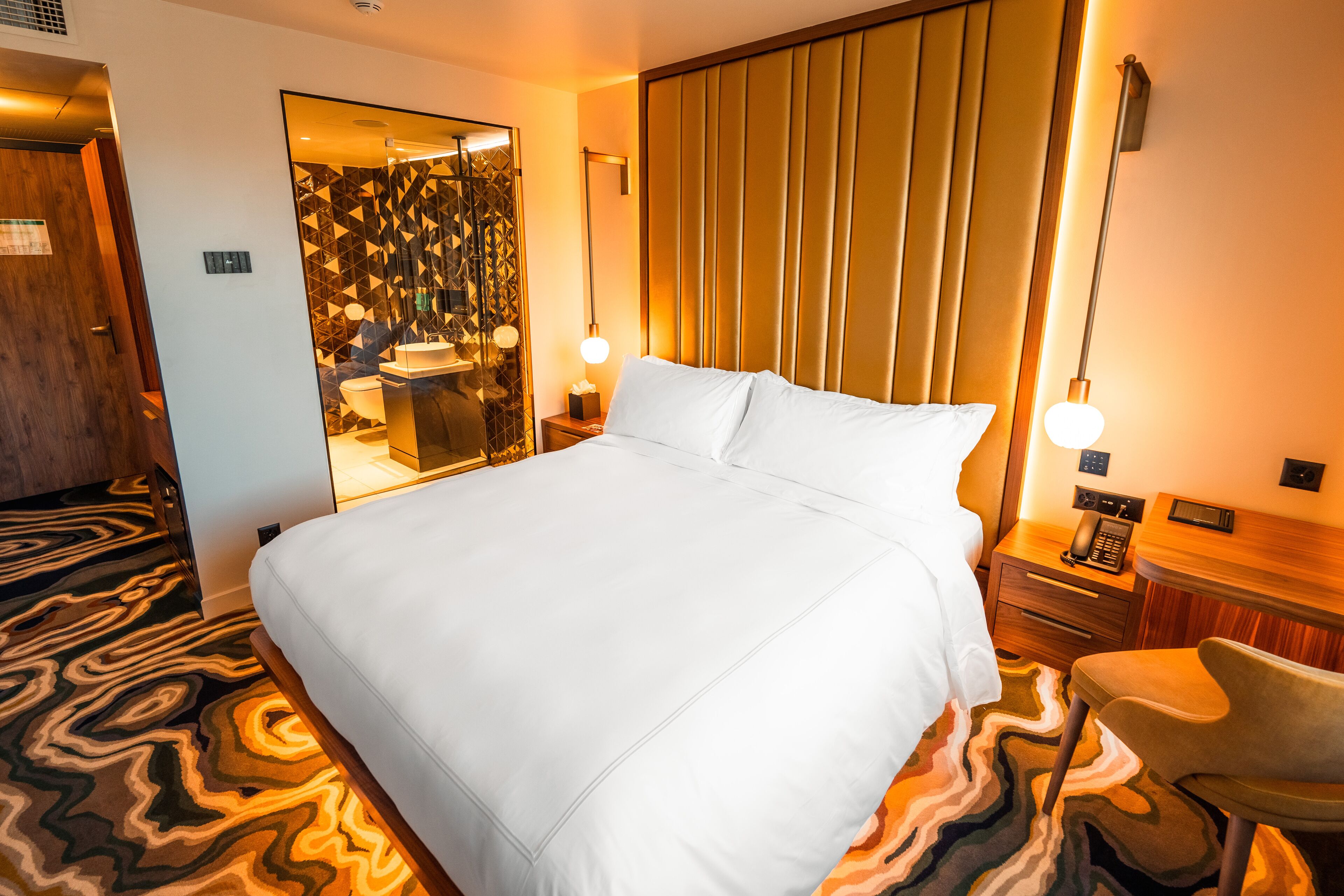 Una camera d'albergo con un letto grande, una sedia, un comodino con una lampada e uno specchio.