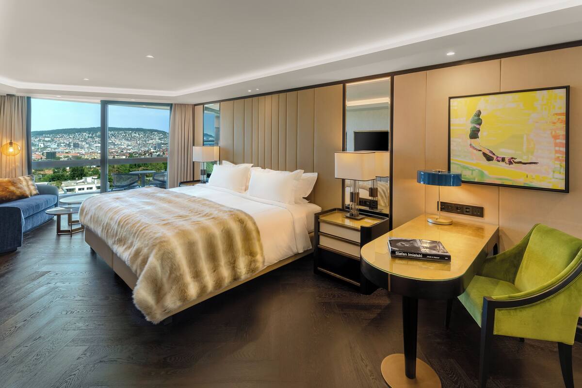 A hotel room with a large bed, a desk, a chair, and a view of a cityscape.
