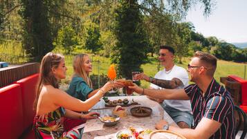 Un groupe de personnes est assis à une table à l’extérieur, devant de la nourriture et des boissons, et est entouré de verdure.
