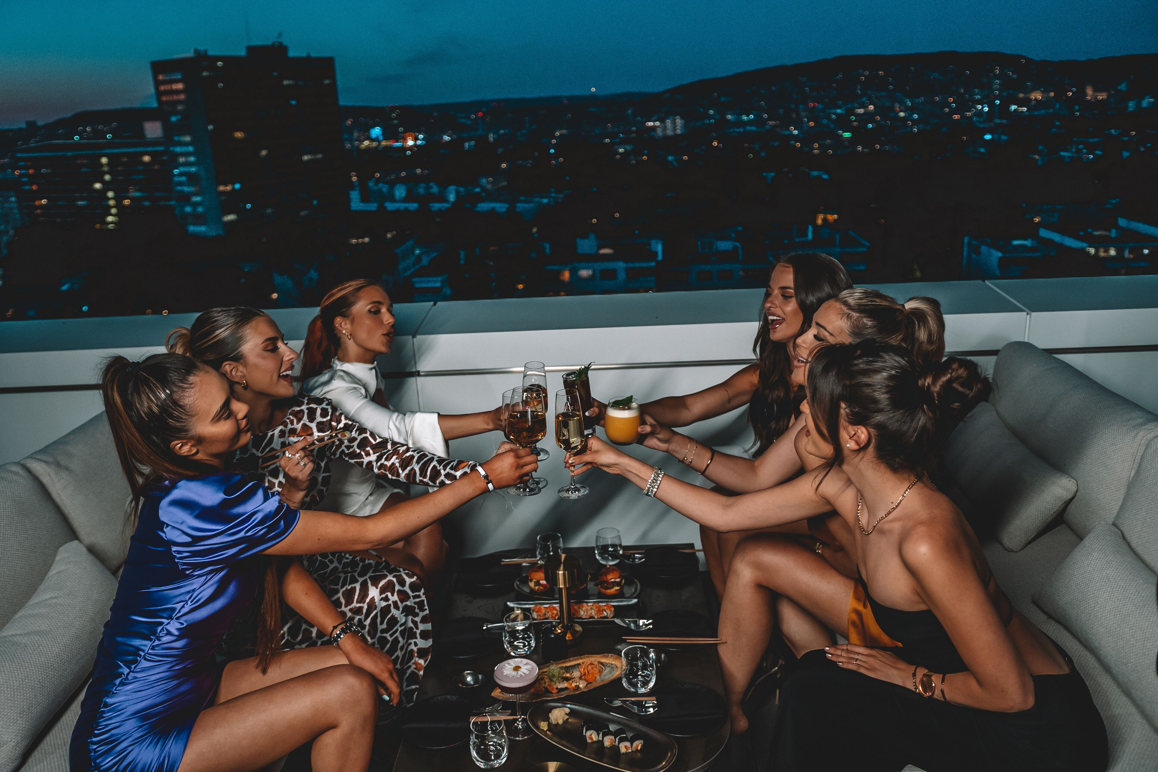 Un groupe de personnes trinquant avec des boissons autour d’une table où sont disposés de la nourriture et des breuvages, le tout sur fond de paysage urbain.