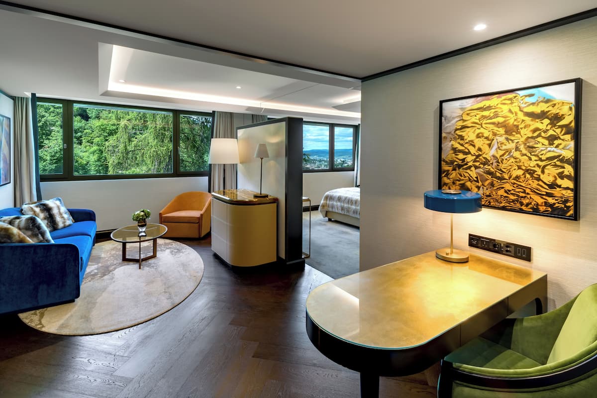 A modern hotel room with a large window, a blue sofa, a green chair, a small round table, and a lamp.