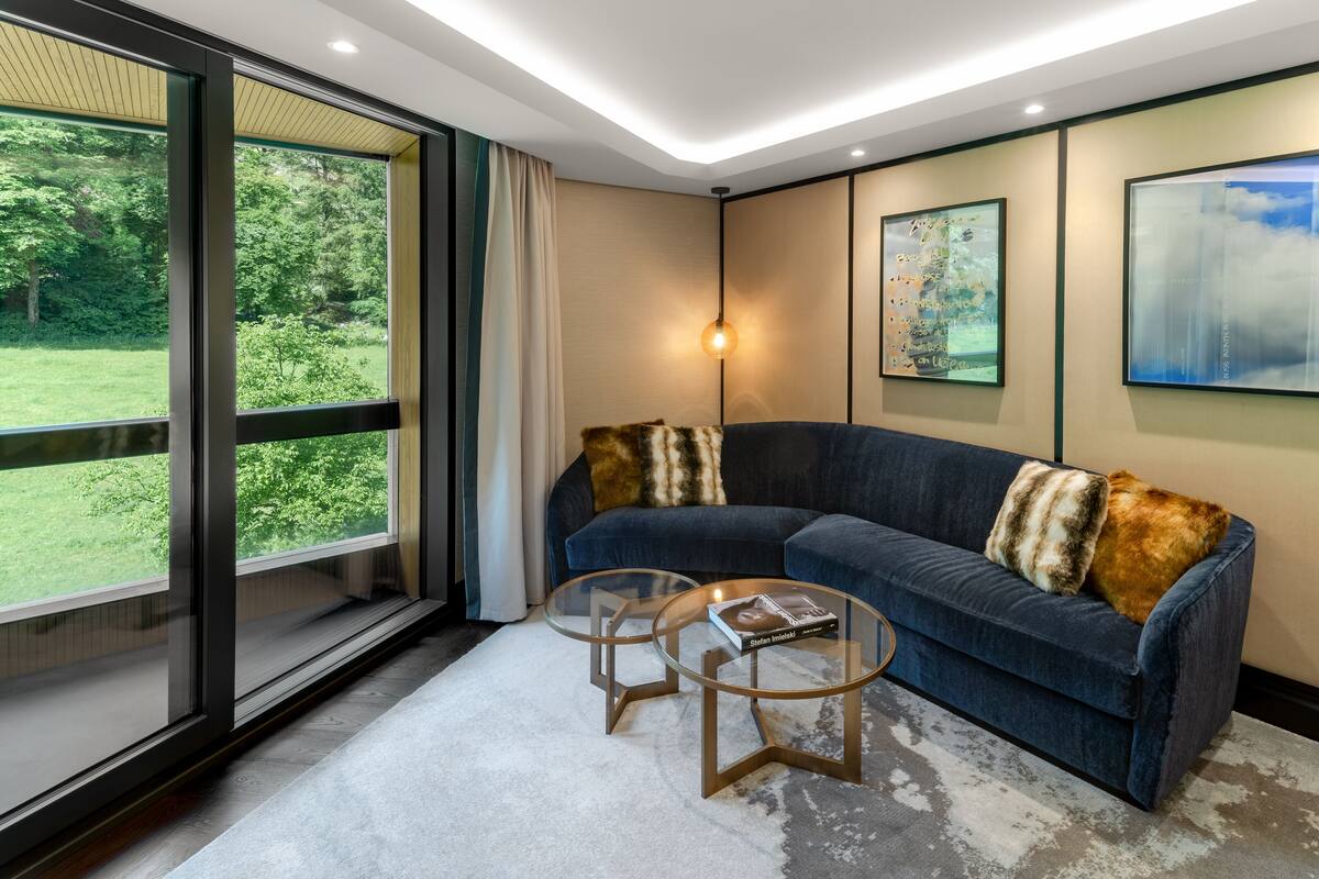A modern living room with a dark blue sofa, two glass coffee tables, and a large window offering a view of a green landscape.