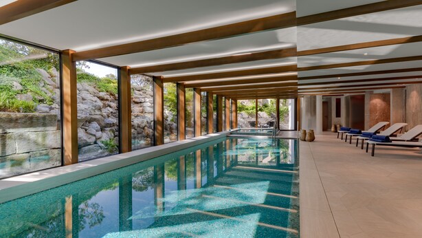 Una piscina interior moderna con paredes de vidrio transparente, vigas de madera y sillones de descanso.
