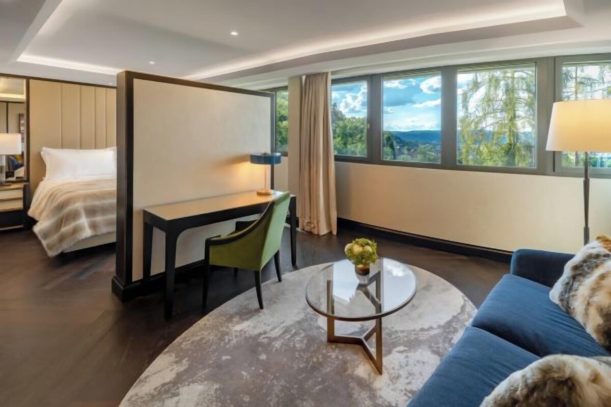 A modern hotel room with a bed, a desk, a round coffee table with a plant, a blue sofa, and large windows offering a view of greenery.