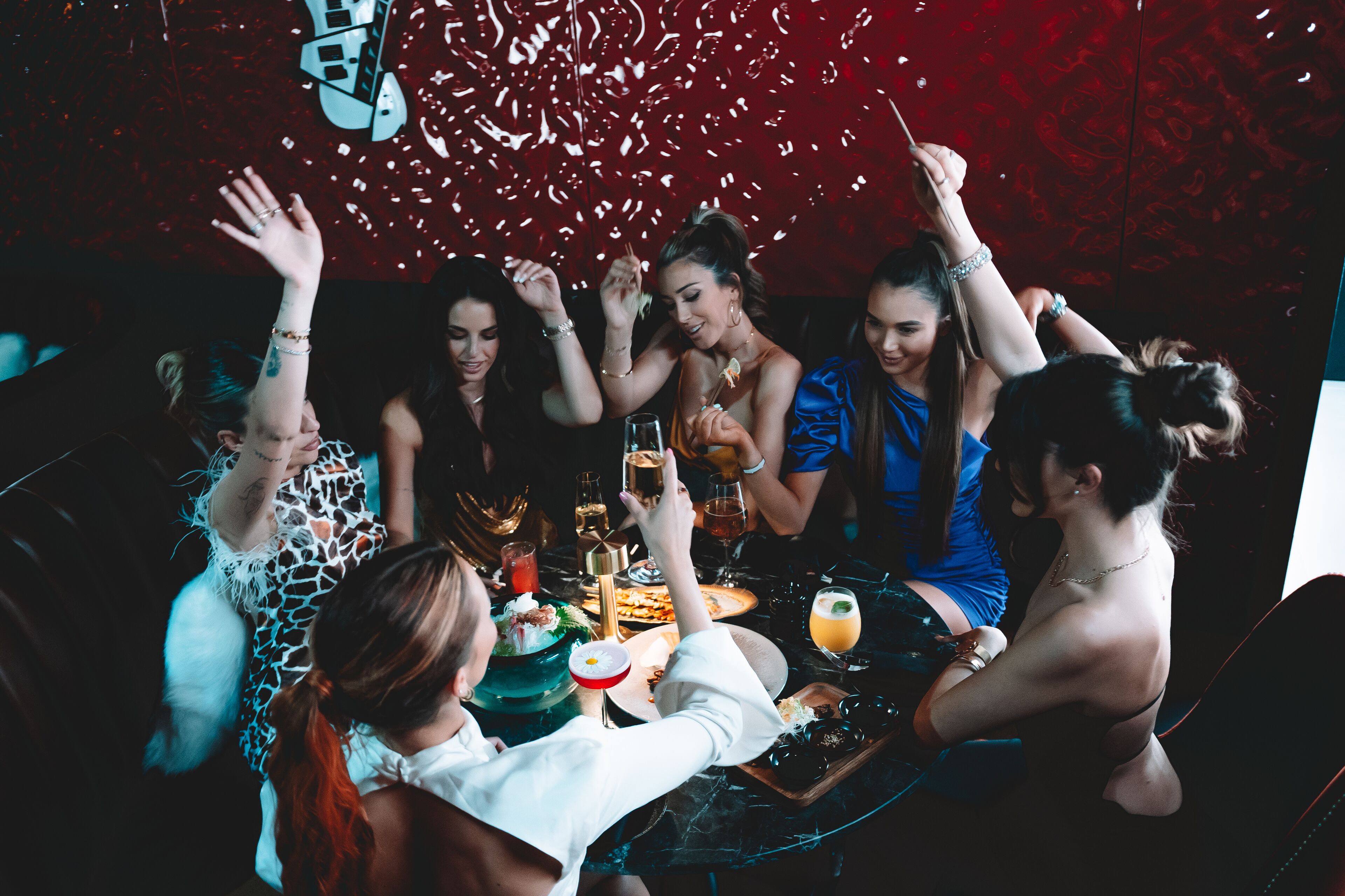 A group of people at a table with drinks and food, in a room with a red patterned wall.