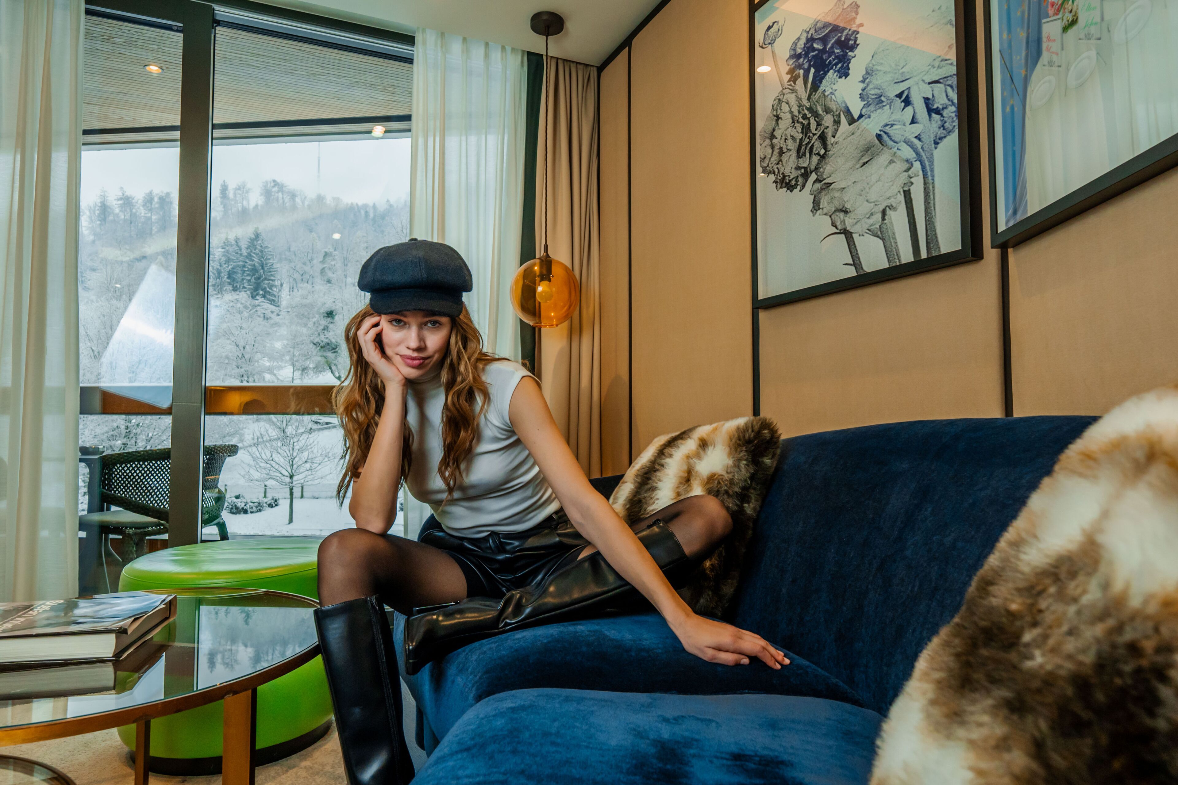Una mujer sentada en un sofá azul en una habitación con un ventanal grande, una silla verde y una mesa con un libro.