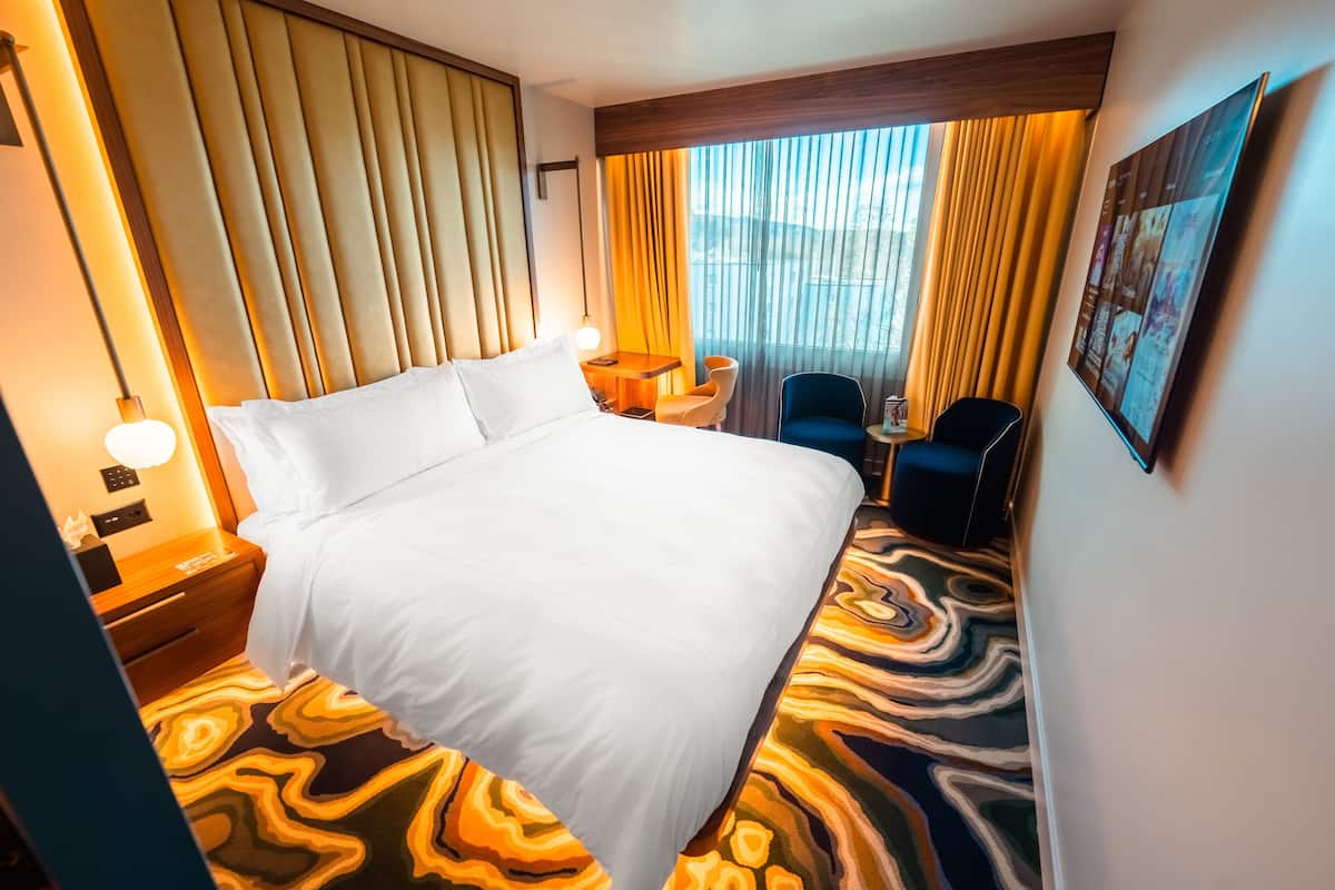 A hotel room with a large bed, a desk with a chair, a small table with a chair, and a large window with curtains.