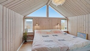 Une chambre avec un plafond en pente, des murs en bois, un lit recouvert d’une couvre-lit à motifs, deux lampes de chevet et une grande fenêtre.